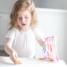 Load image into Gallery viewer, A young girl grabs snacks from a colorful Bumkins Reusable Snack Bag, Large 2-Pack: Watercolor & Brush Strokes at a white table.