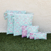 Load image into Gallery viewer, Bumkins Wet Bag: Rainbows sits on grass against a textured beige wall among other matching waterproof bags.