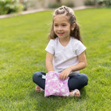 A girl with long hair sits on grass, smiling while holding a Bumkins Reusable Snack Bag, Large 2-Pack: Rainbows & Unicorns.