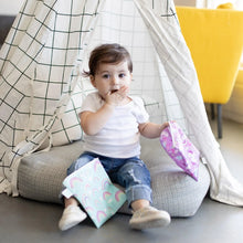 Load image into Gallery viewer, A toddler covers their mouth, sitting in a checkered fabric tent and holding Bumkins Reusable Snack Bag, Large 2-Pack: Rainbows & Unicorns.