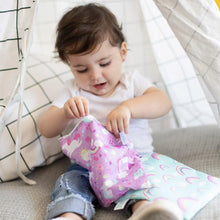 Load image into Gallery viewer, A toddler in jeans opens a Bumkins Reusable Snack Bag, Large 2-Pack: Rainbows & Unicorns while sitting inside a cloth tent.