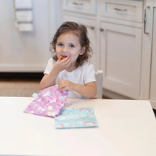 Load image into Gallery viewer, A young girl in a white shirt enjoys snacks from Bumkins Reusable Snack Bag, Large 2-Pack: Rainbows & Unicorns at a white table.
