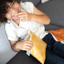 Load image into Gallery viewer, A child in a white shirt and jeans laughs on a couch, holding Bumkins Reusable Snack Bag, Large 2-Pack: Sunshine and Grounded.