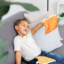 Load image into Gallery viewer, A child sits on a couch, smiling as he holds a Bumkins Reusable Snack Bag, Large 2-Pack: Sunshine and Grounded, with the other bag on his lap.