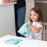 A girl holds a Bumkins Reusable Snack Bag, Large 2-Pack: Ocean Life & Whale Tail while another sits nearby on a white table.