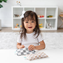 Load image into Gallery viewer, A smiling girl with a white bow holds a Bumkins Reusable Snack Bag, Small 2 Pack: Double Check, at a white table with a shelf behind her.