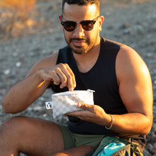 Load image into Gallery viewer, A man in sunglasses reaches into a Roamio by Bumkins Reusable Snack Bag, Small 2 Pack: Rich & Boujee outdoors in golden sunlight.
