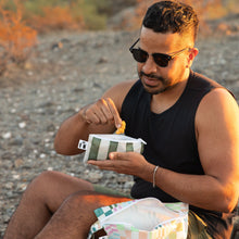 Load image into Gallery viewer, A man wearing sunglasses enjoys snacks from a Roamio by Bumkins Reusable Snack Bag, Small 2 Pack: Motley Club while sitting on rocky ground.