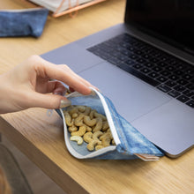 Load image into Gallery viewer, A hand opens a Roamio by Bumkins Reusable Snack Bag, Small 2 Pack: Denim on Denim next to a laptop, revealing cashews inside.