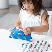 Load image into Gallery viewer, A young girl in a white dress opens Bumkins Reusable Snack Bag, Small 2 Pack: Mickey & Friends MVP at a white table.