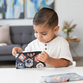 A young boy looks into a Bumkins Reusable Snack Bag, Small 2 Pack: Everyday Is Halloween at a white table in a modern living room.