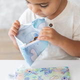A toddler in a white shirt examines snacks in a Bumkins Reusable Snack Bag, Small 2 Pack: Stitch Star-ring Up Trouble at a white table.