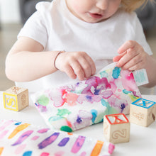 Load image into Gallery viewer, A child in a white shirt unzips a Bumkins Reusable Snack Bag, Small 2-Pack: Watercolor & Brush Strokes, surrounded by blocks on a table.