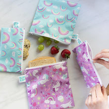 Load image into Gallery viewer, A person zips Bumkins Reusable Snack Bag, Small 2-Pack: Rainbows & Unicorns on a marble surface with other bags and snacks close by.