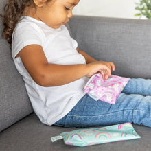 Load image into Gallery viewer, A child in jeans and a white shirt sits with Bumkins Reusable Snack Bag, Small 2-Pack: Rainbows & Unicorns on a gray couch.