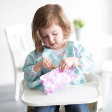 Load image into Gallery viewer, A toddler in a high chair wearing a blue bib enjoys snacks from a Bumkins Reusable Snack Bag, Small 2-Pack: Rainbows & Unicorns.