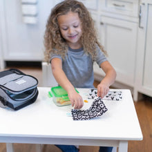 Load image into Gallery viewer, A curly-haired girl zips a Bumkins Reusable Snack Bag, Small 2-Pack: XOXO & Hearts at a white table with a lunch box and food container.
