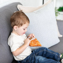 Load image into Gallery viewer, A young boy sits on a gray couch, peeking inside an orange Bumkins Reusable Snack Bag, Small 2-Pack: Sunshine and Grounded.