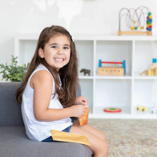 Load image into Gallery viewer, A smiling girl sits on a sofa with Bumkins Reusable Snack Bag, Small 2-Pack: Sunshine and Grounded. Shelves with toys are in the background.