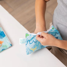 Load image into Gallery viewer, Close-up of a child reaching into a Bumkins Reusable Snack Bag, Small 2-Pack: Ocean Life & Whale Tail at a white table.