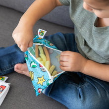 Load image into Gallery viewer, A child on a gray couch opens a blue Bumkins Reusable Snack Bag, Small 2-Pack: Super Mario™, revealing veggie sticks inside.