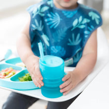 Load image into Gallery viewer, A toddler in a blue bib holds a Bumkins Silicone Straw Cup with Lid: Blue, sitting in a high chair beside a plate of varied foods.
