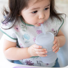 Load image into Gallery viewer, A child uses a Bumkins Silicone Straw Cup with Lid: Gray, honing their drinking skills while wearing a floral bib.