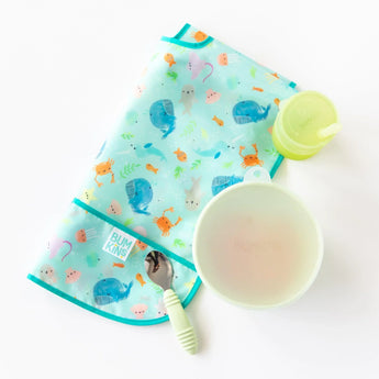 Bumkins Silicone Straw Cup with Lid: Sage sits near a folded ocean-themed bib, a green bowl with lid, & a green spoon on a white background.