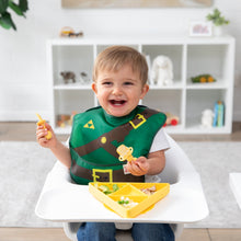 Load image into Gallery viewer, A boy wearing a Bumkins Caped SuperBib®: The Legend of Zelda™ Link smiles in a high chair, holding yellow utensils by a matching plate.