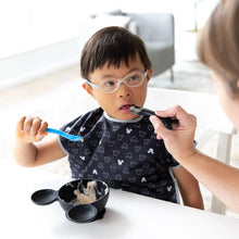 Load image into Gallery viewer, A child with glasses self-feeds from a bowl using a Bumkins Silicone Dipping Spoons: Mickey Mouse (Black and Blue), assisted by an adult.