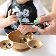 Load image into Gallery viewer, An adult helps feed a child from a gold bowl of blueberries with a Bumkins Silicone Dipping Spoons: Mickey Mouse (Classic Black).