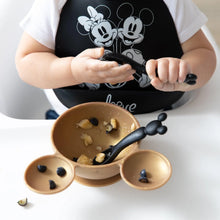 Load image into Gallery viewer, A child in a black bib holds a Bumkins Silicone Dipping Spoons: Mickey Mouse (Classic Black), with a gold bowl of food on a high chair tray.
