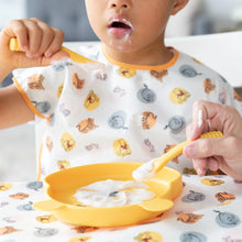 Load image into Gallery viewer, A young child in a colorful bib eats from a plate with Bumkins Silicone Dipping Spoons: Winnie the Pooh, while an adult assists.