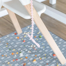 Load image into Gallery viewer, A tether attached to a high chair holds a Bumkins Silicone Dipping Spoons 3 Pack: Lollipop, dangling above a floor mat covered with food.