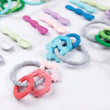 Bumkins Silicone Teething Charms: Pink lays next to other teething toys and silicone spoons on a white marble background.