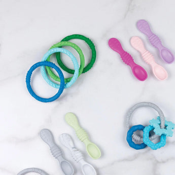 Bumkins Silicone Teething Rings 4 Pack: Summer lays near other teething toys and silicone spoons on a white marble background.