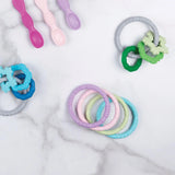 Bumkins Silicone Teething Rings 4 Pack: Spring rests among other teething toys and silicone spoons on a marble background.