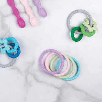 Bumkins Silicone Teething Rings 4 Pack: Spring rests among other teething toys and silicone spoons on a marble background.