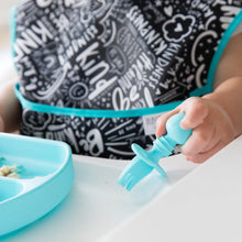 Load image into Gallery viewer, Close-up of a baby in a black & white bib holding a Bumkins Silicone Chewtensils®: Blue next to a blue divided plate.