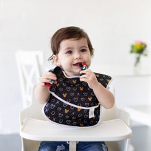 Load image into Gallery viewer, A baby in a high chair wearing a black Bumkins SuperBib® 3 Pack: Love, Minnie smiles while holding the Silicone Chewtensils®: Minnie Mouse.