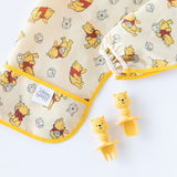 Close-up of a folded Bumkins Sleeved Bib: Winnie Loves Hunny on a white background, with Winnie the Pooh-shaped utensils nearby.