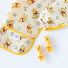 Load image into Gallery viewer, Close-up of a folded Bumkins Sleeved Bib: Winnie Loves Hunny on a white background, with Winnie the Pooh-shaped utensils nearby.