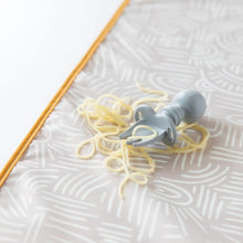 Load image into Gallery viewer, Close-up of a Bumkins Silicone Chewtensils®: Gray with spaghetti on a beige waterproof mat on a white background.