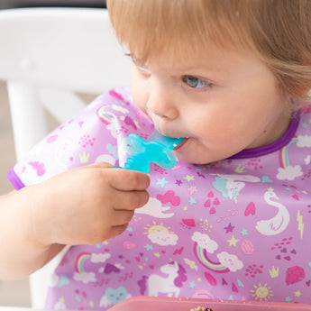 Close-up of a child in a purple unicorn bib chewing a Bumkins Silicone Chewtensils®: Blue Jelly.