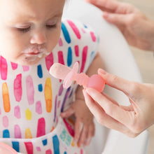Load image into Gallery viewer, An adult hand uses a Bumkins Silicone Chewtensils®: Pink Jelly to feed a child with a yogurt-covered mouth in a colorful bib.
