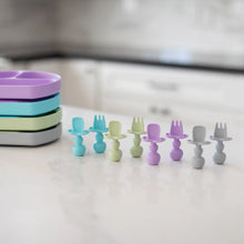 Load image into Gallery viewer, Bumkins Silicone Chewtensils®: Lavender are arranged with other silicone utensils near a stack of divided plates on a marble kitchen counter