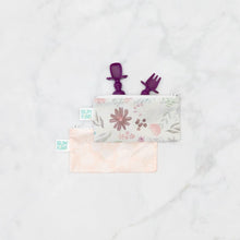 Load image into Gallery viewer, Bumkins Silicone Chewtensils®: Purple peeks from a floral zippered pouch, with another pink pouch underneath on a marble background.