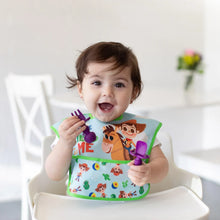 Load image into Gallery viewer, A smiling toddler in a high chair, wearing a colorful bib, holds Bumkins Silicone Chewtensils®: Purple in a bright room.
