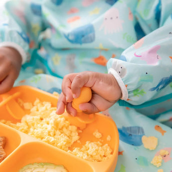 Close-up of a child in a blue sea-themed bib enjoying eggs from an orange divided plate using Bumkins Silicone Chewtensils®: Tangerine.