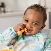 Load image into Gallery viewer, A smiling baby in a high chair, wearing a a dinosaur bib, chews a Bumkins Silicone Chewtensils®: Tangerine.